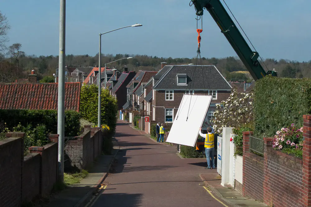 Mobiele kraan in een smalle straat tilt een witte dakkapel boven een rijtjeshuis
