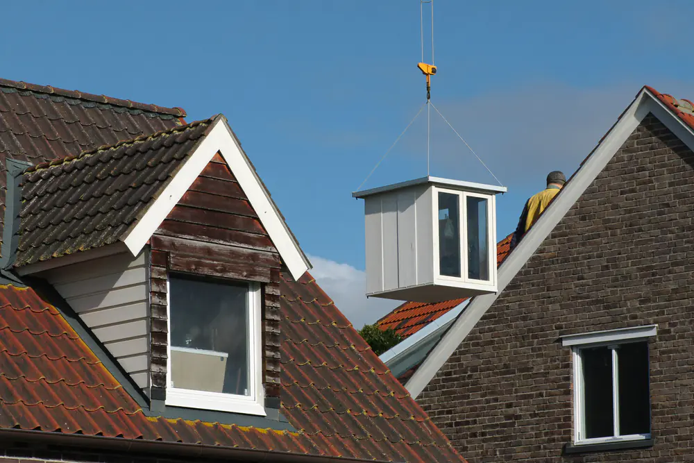 Een oude houten dakkapel naast een nieuwe dakkapel die met een kraan wordt geplaatst
